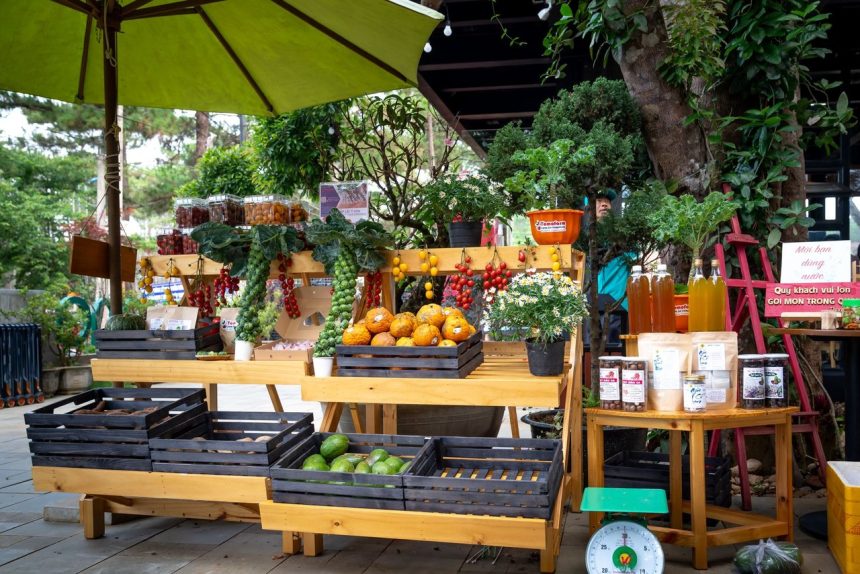 Banca de mercado ao ar livre com frutas, verduras e outros produtos sob um guarda-sol verde.