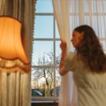 Mulher observa o entardecer pela janela enquanto afasta as cortinas de um ambiente aconchegante.