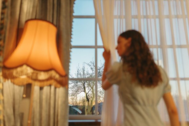Mulher observa o entardecer pela janela enquanto afasta as cortinas de um ambiente aconchegante.