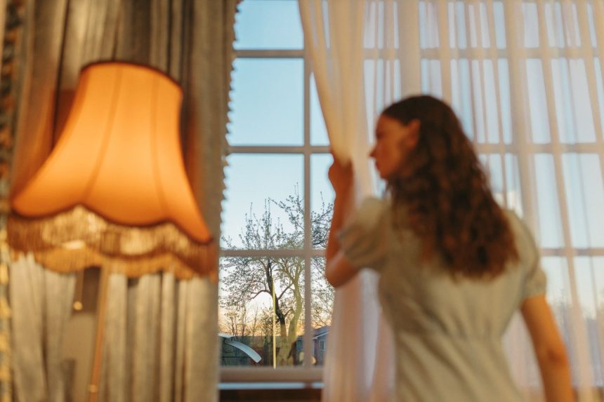 Mulher observa o entardecer pela janela enquanto afasta as cortinas de um ambiente aconchegante.