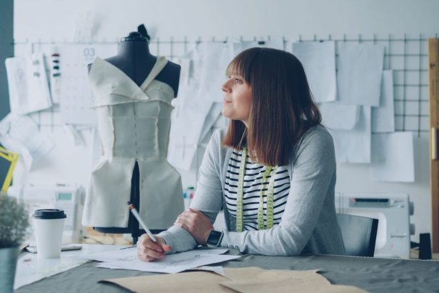 Designer planeja novas criações em um estúdio de moda, cercado por materiais e referências de trabalho.