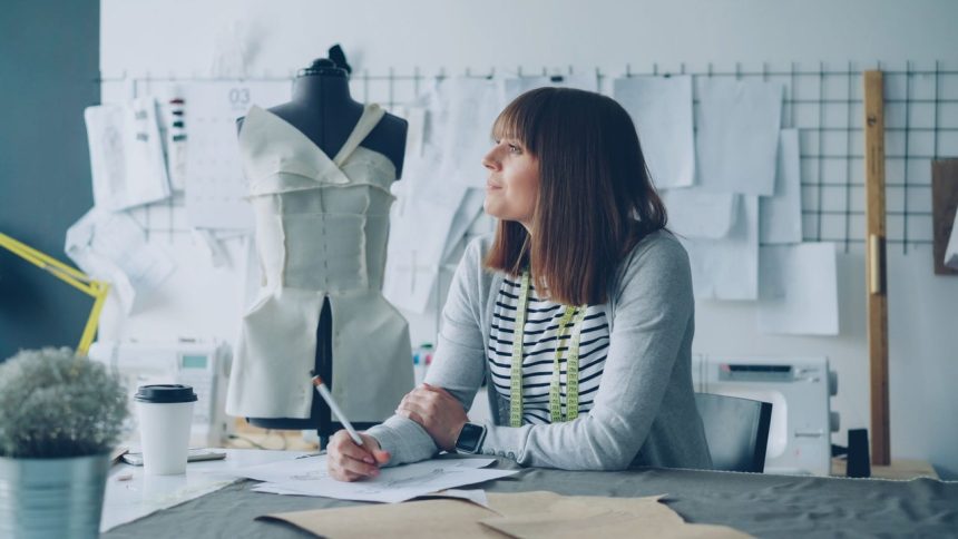 Designer planeja novas criações em um estúdio de moda, cercado por materiais e referências de trabalho.
