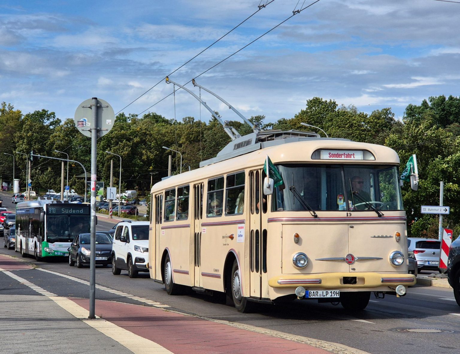 Trolebus circula por uma rua urbana arborizada, entre veículos e edifícios.