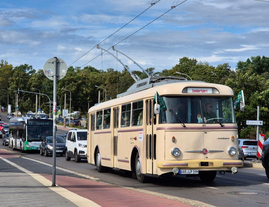 Trolebus circula por uma rua urbana arborizada, entre veículos e edifícios.